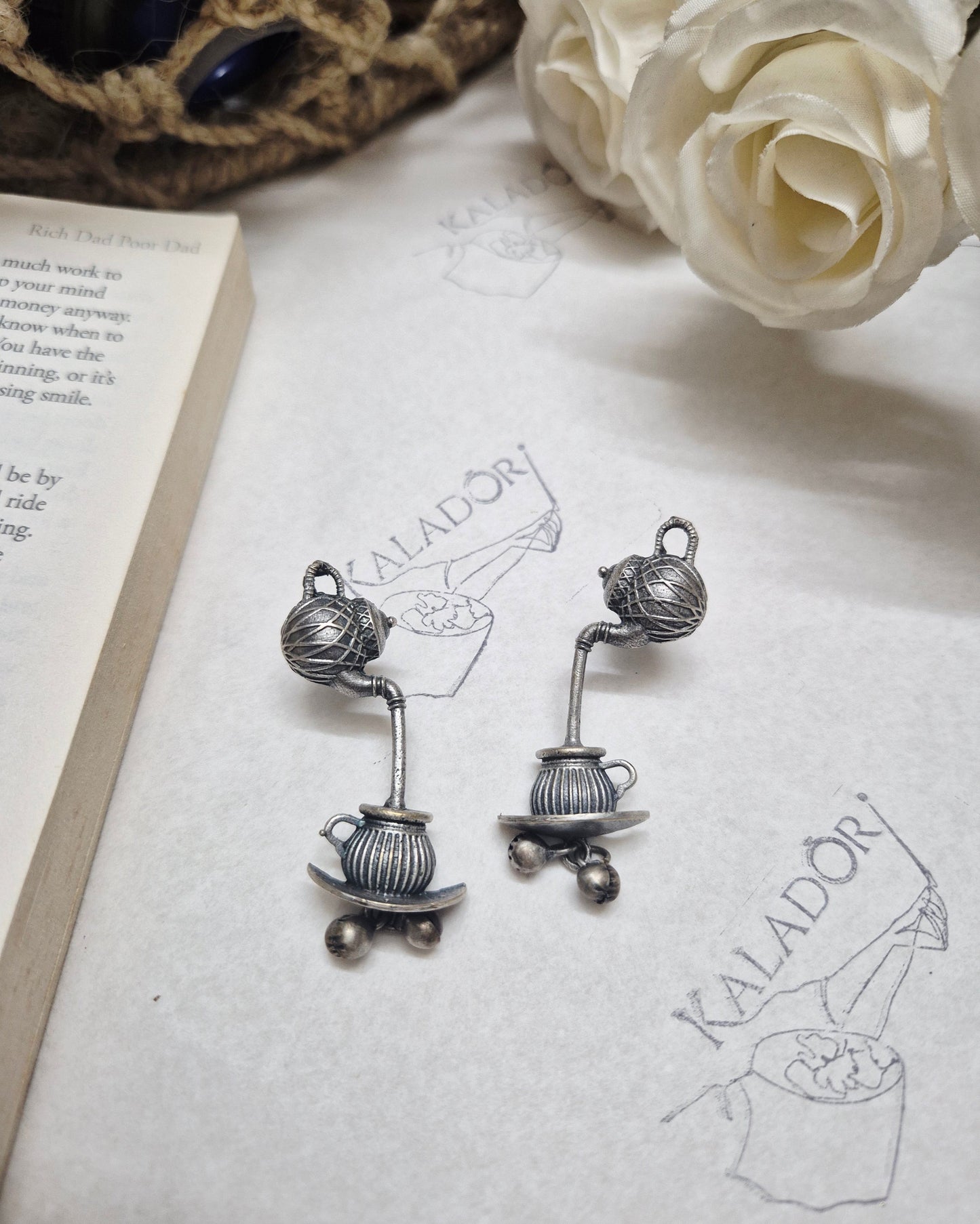 Pair of silver earrings on a white surface with a flower and book in the background.