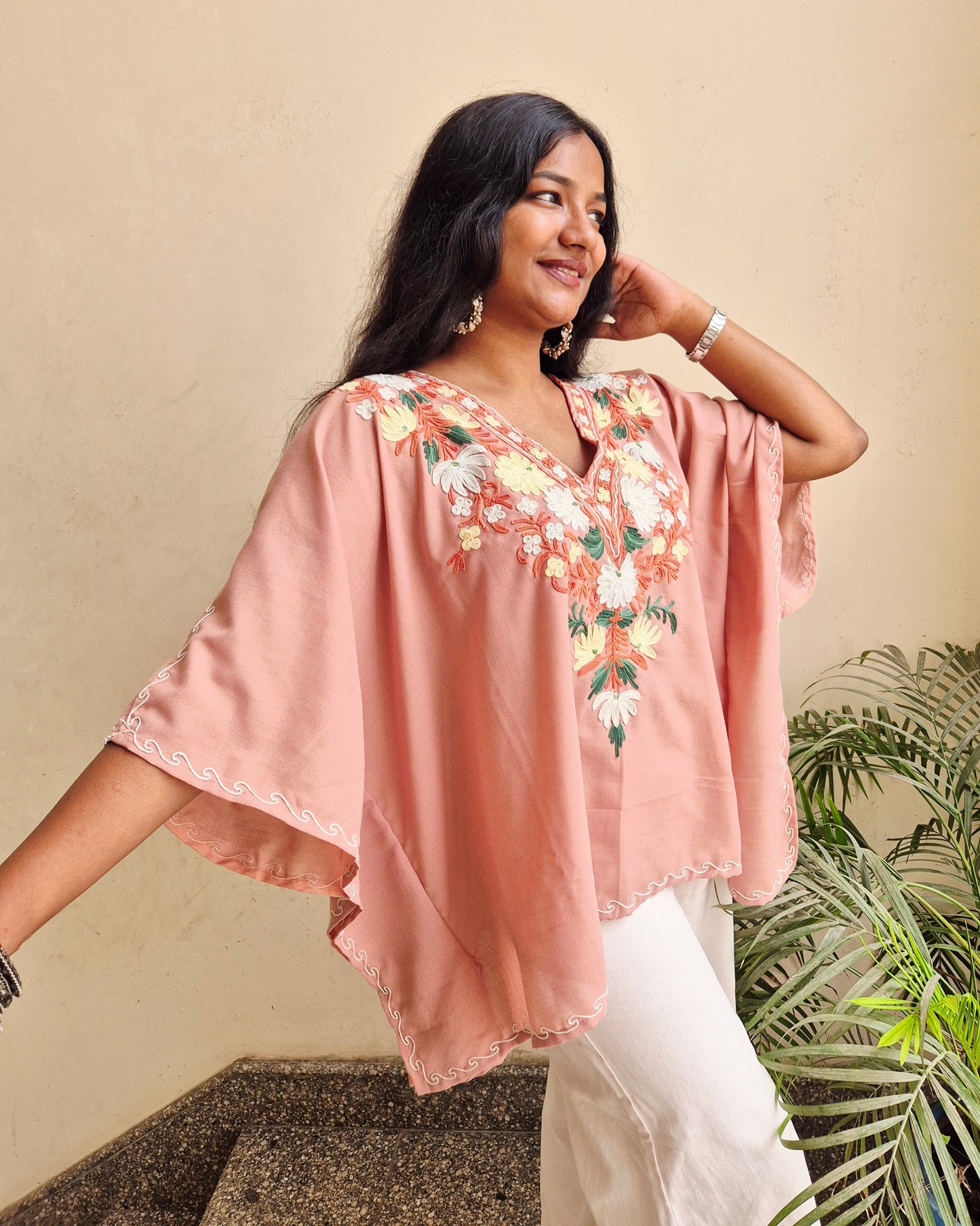 Woman wearing a pink floral embroidered top and white pants against a beige wall with green plants.