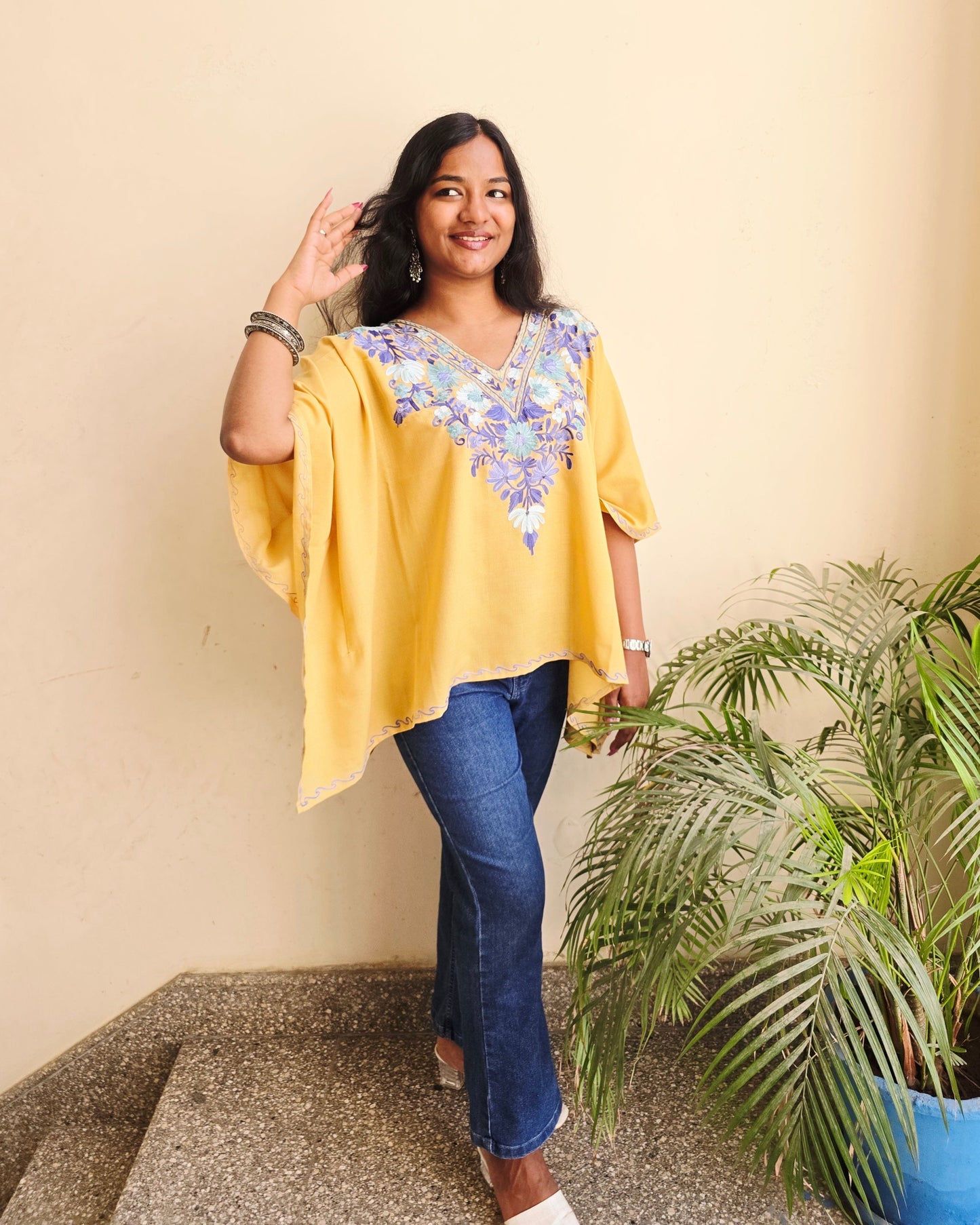 Person wearing a yellow top and blue jeans standing next to a plant on a beige wall.