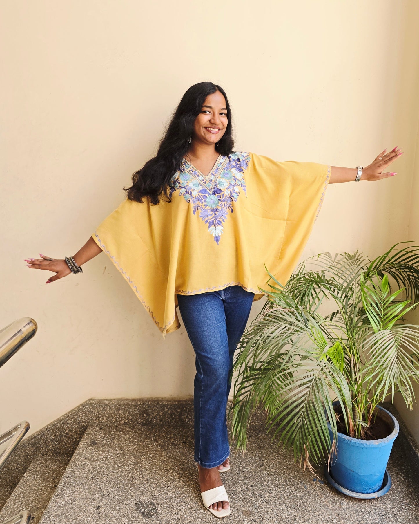 Woman in yellow top and blue pants standing on a staircase with plants around