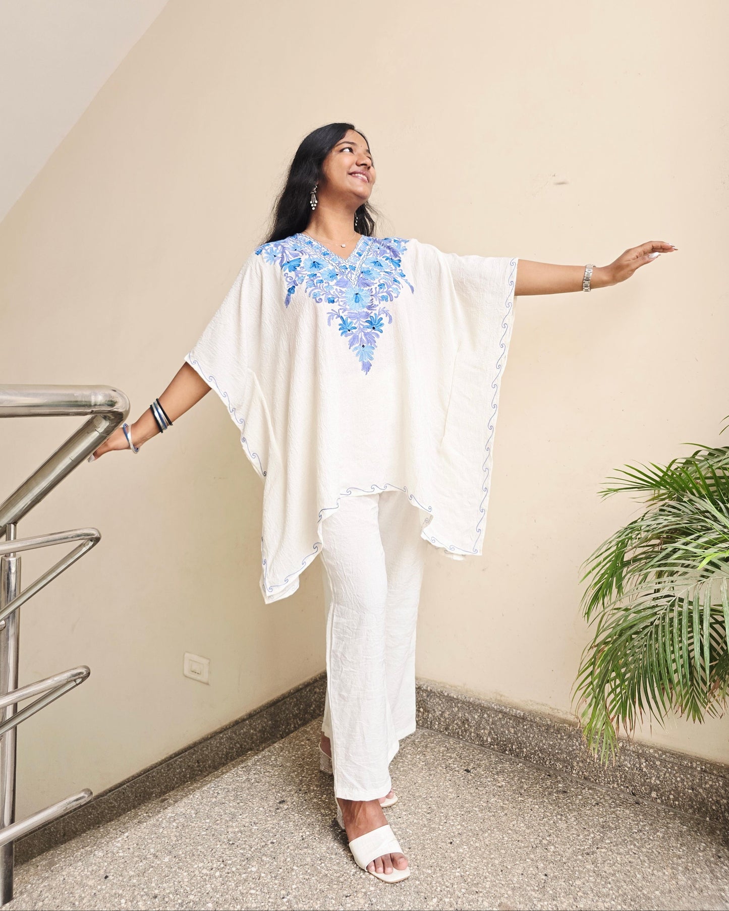 Person in a white outfit with floral design standing on a railing indoors.