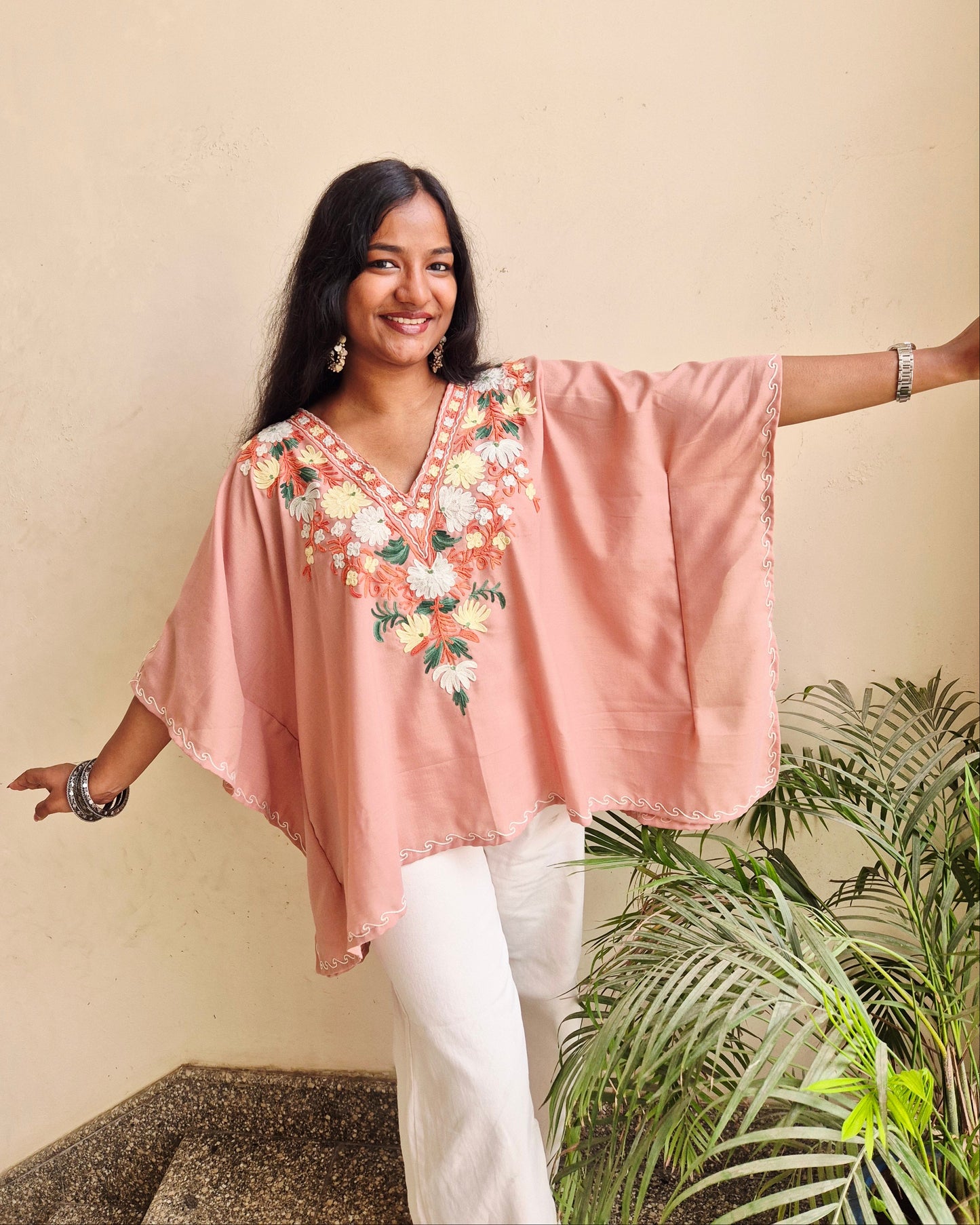 Woman wearing a pink embroidered top with a beige wall and green plant in the background