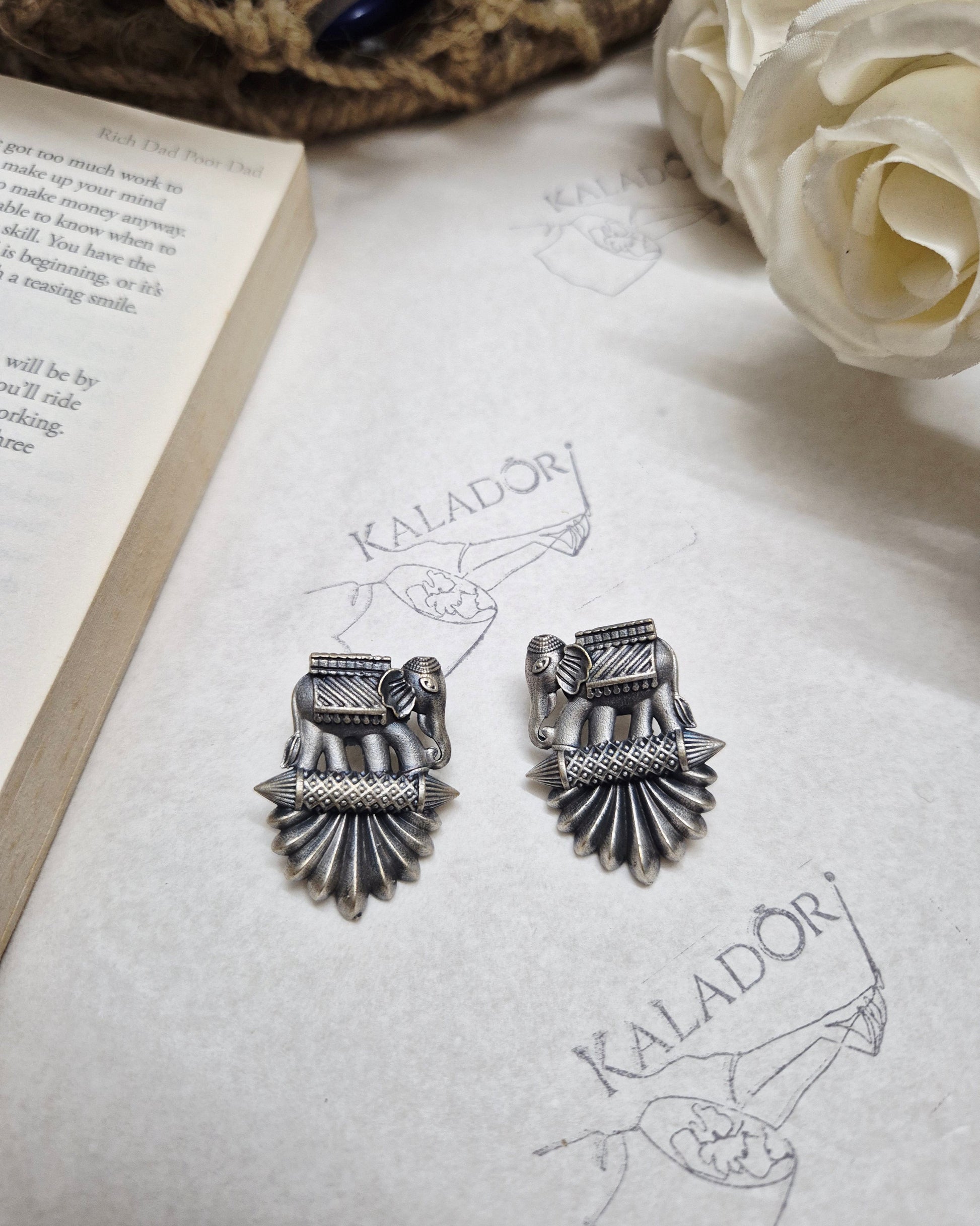 Silver earrings on a piece of paper with 'Kalador' branding, next to a rose and book.
