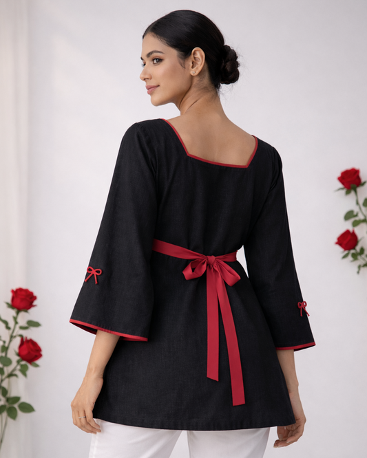 Woman wearing a black top with red trim and a red belt, standing against a white background with red flowers.