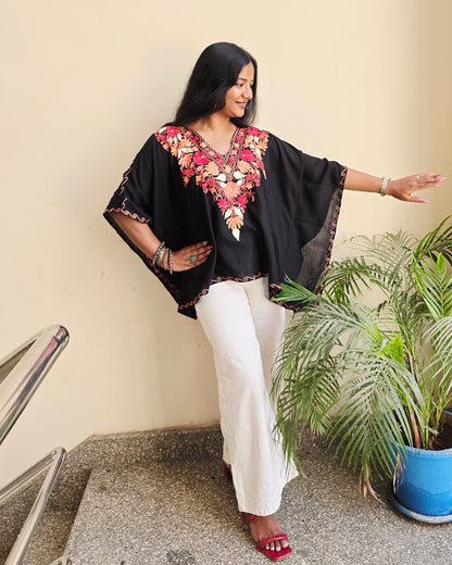 Person in a black outfit with floral patterns standing next to a plant indoors.