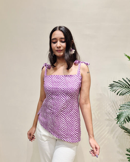 Woman wearing a purple striped short kurti and white pants against a beige wall with green leaves.