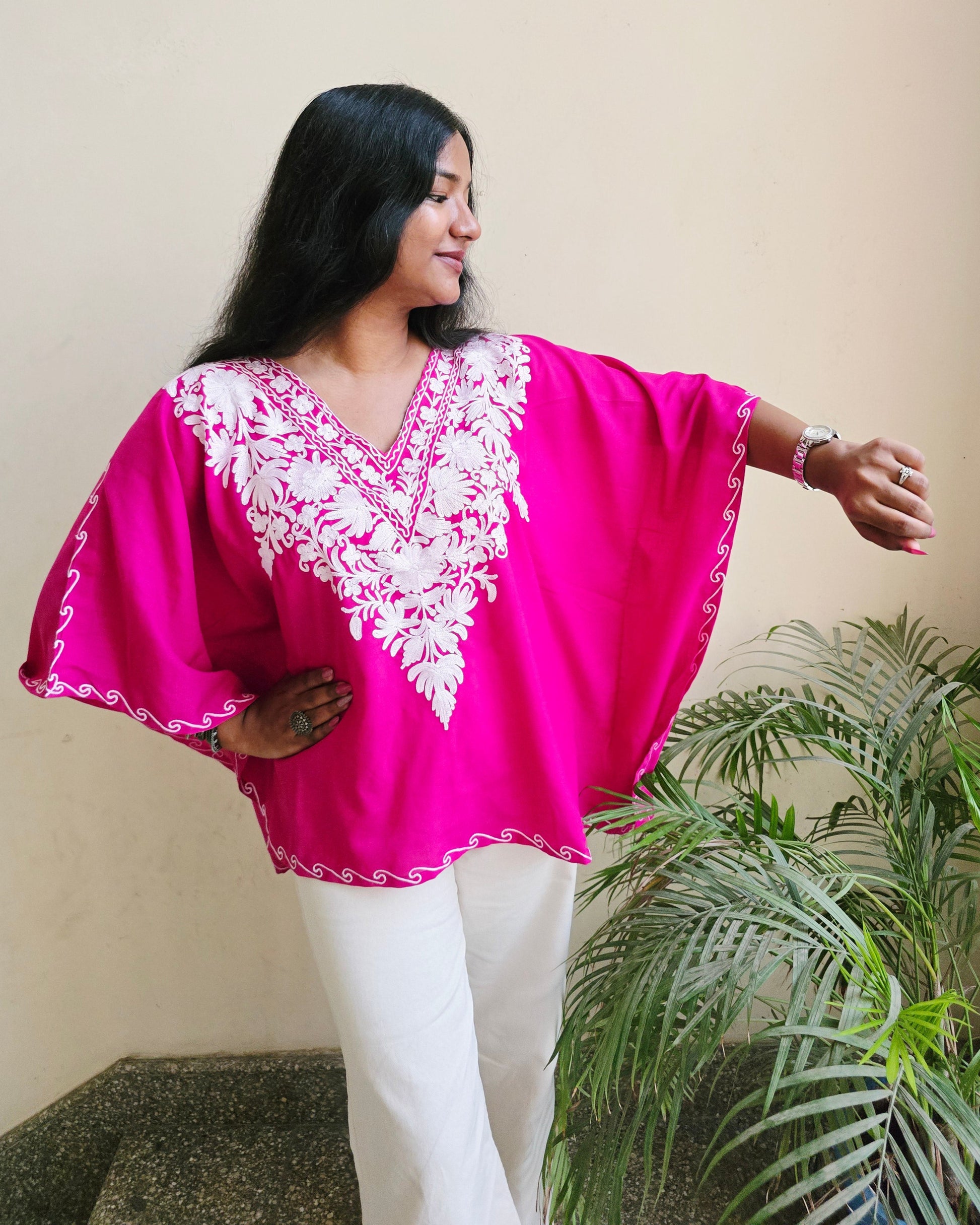 Woman wearing a bright pink traditional outfit with white embroidery, standing against a plain wall with a plant in the background.