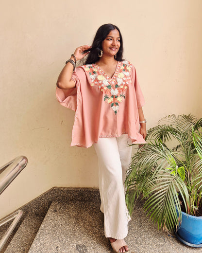 Woman in a pink dress with floral embroidery standing indoors next to a plant.